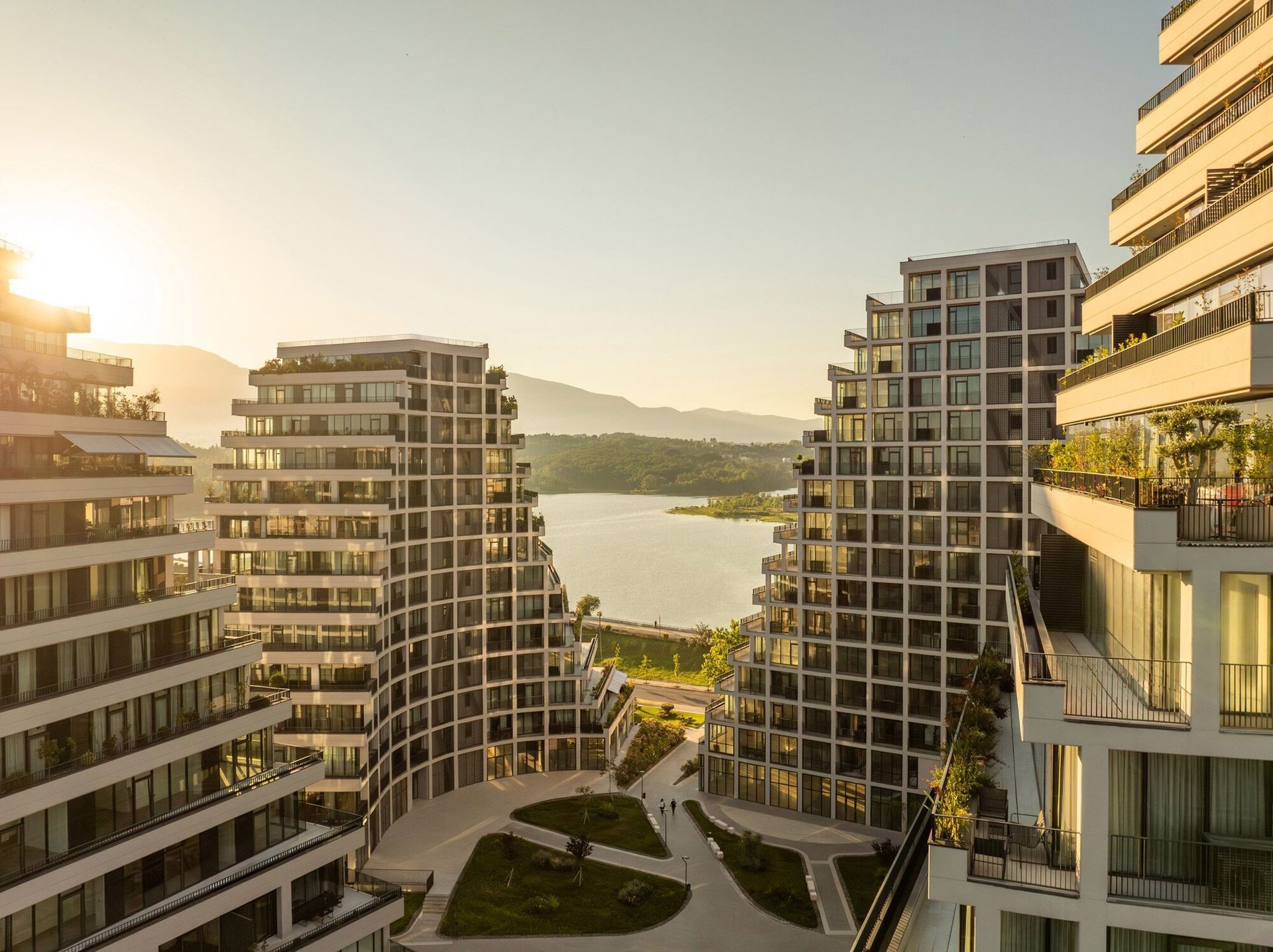 Modern residential buildings surrounding a scenic lake at sunset with forested hills in the background
